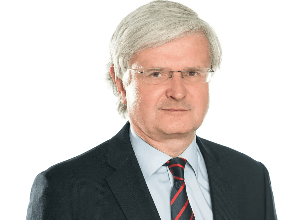 Man with gray hair and glasses wearing a suit and striped tie, posing against a white background.