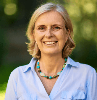 Smiling person with short blond hair wearing a blue shirt and turquoise necklace, standing outdoors.