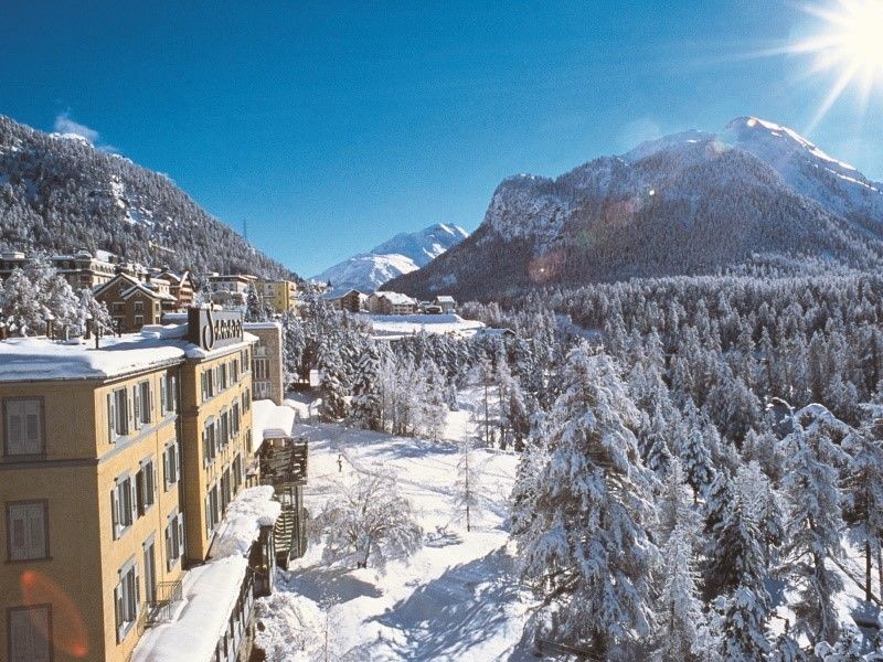 Snow-covered mountain village with buildings and pine trees under a bright blue sky and sun.