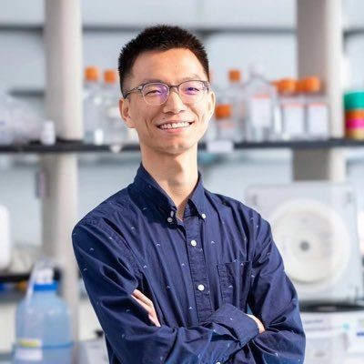 Person wearing glasses and a navy blue shirt, smiling with arms crossed in a laboratory setting.