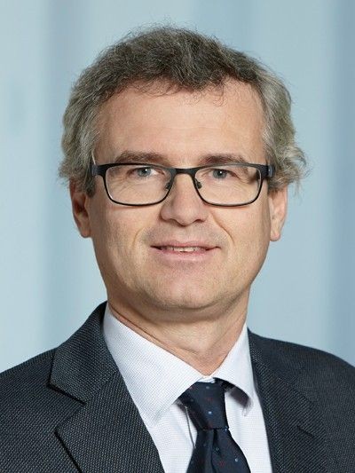 Man in a suit and tie with glasses, gray hair, and a light blue background.