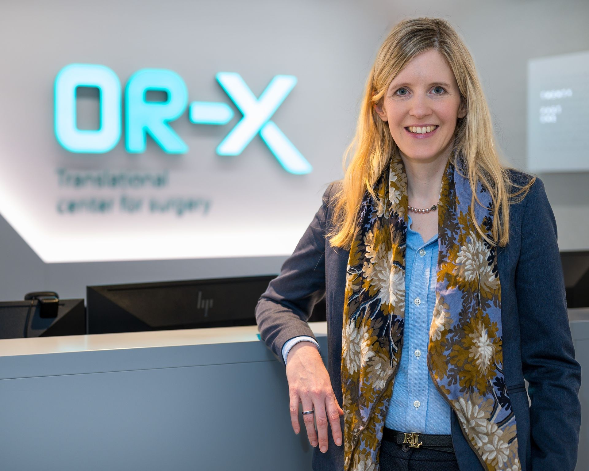 Person standing at a reception desk with a blue and green OR-X logo in the background.