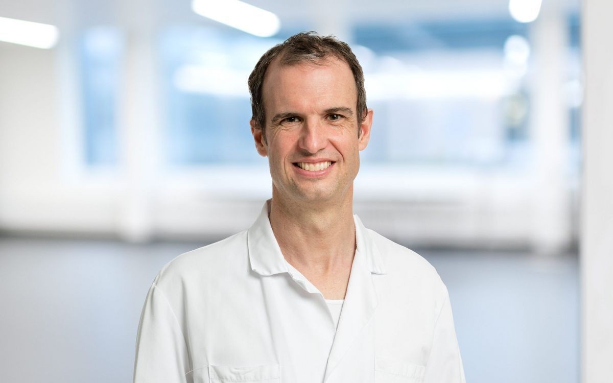 Smiling man in a white lab coat standing in a brightly lit, blurred background.