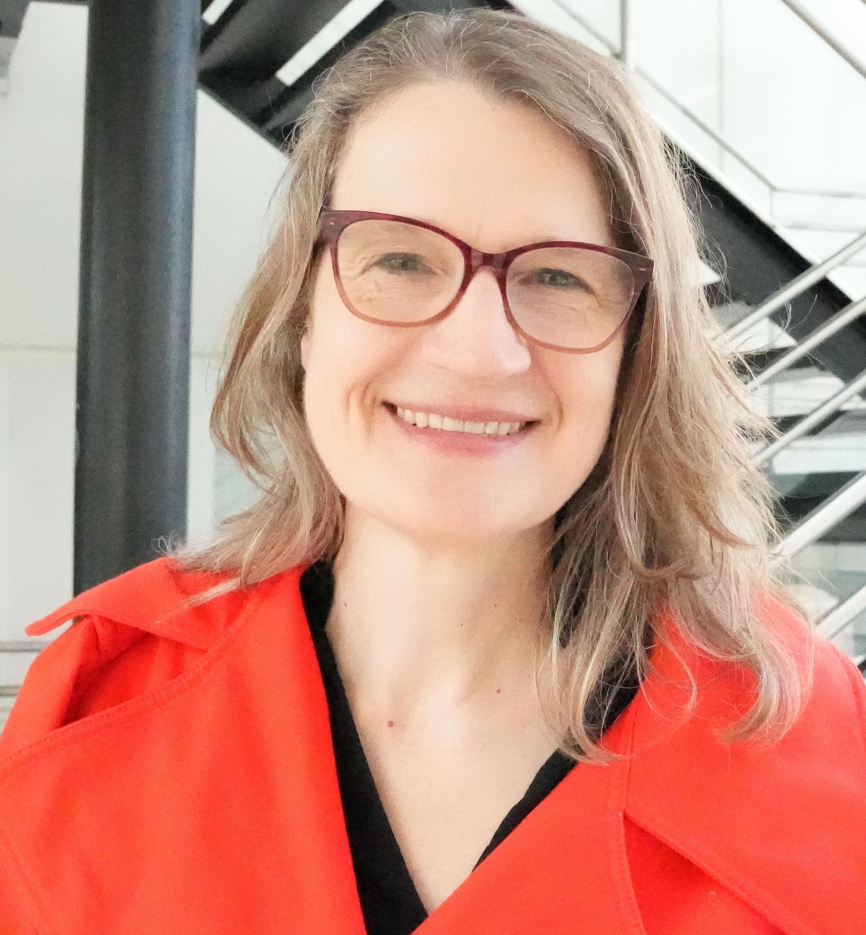 Person with glasses and red jacket, smiling indoors near a staircase.