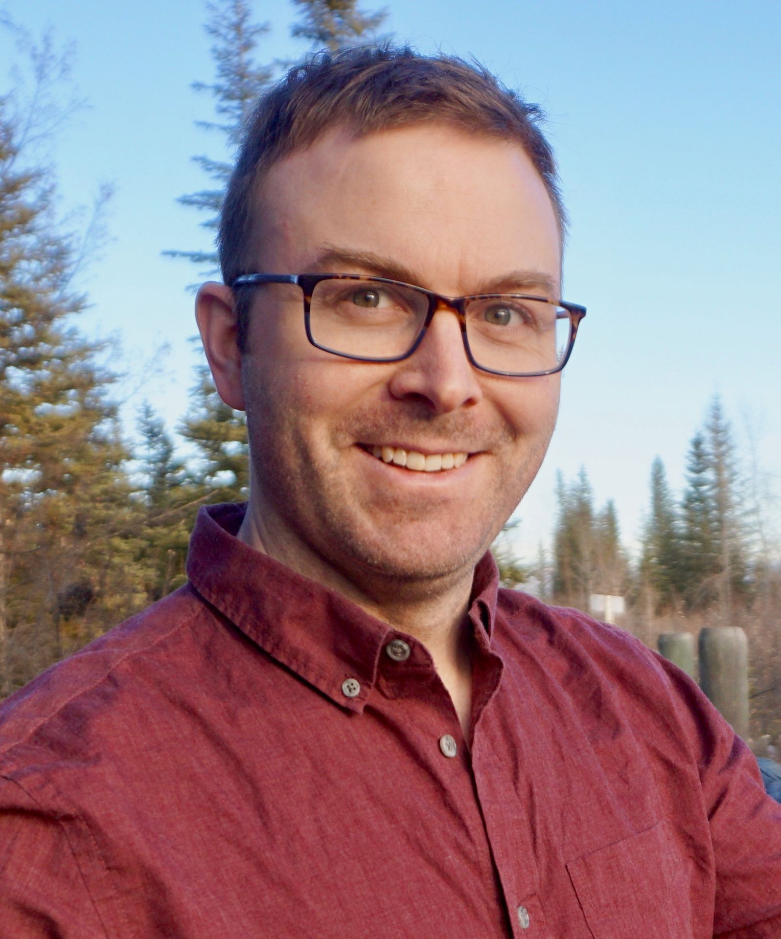 Smiling person with glasses wearing a red shirt, standing outdoors with trees in the background.