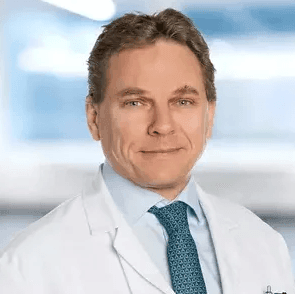 Smiling man wearing a white medical coat and blue tie, standing against a blurred background.