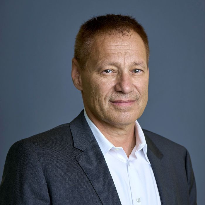 Person wearing a dark suit jacket and white shirt against a plain background.