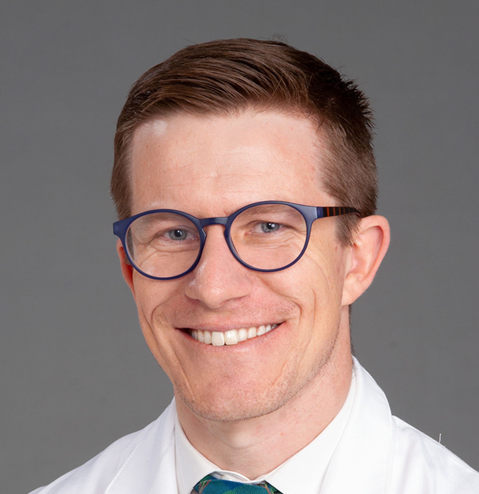 Person with short hair and glasses smiling, wearing a white coat and tie against a gray background.